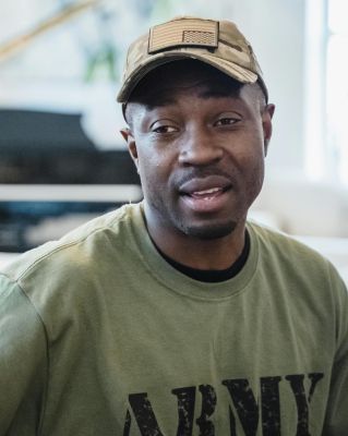 Relaxed portrait of man in army t-shirt and cap sitting in modern living room with piano backdrop.